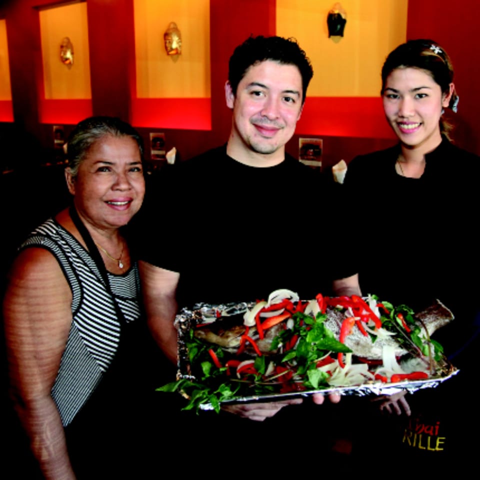 Three Pattaya Thai staff members proudly present a whole steamed fish garnished with herbs and vegetables.
