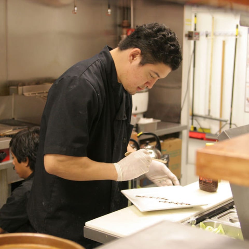 A Pattaya Thai chef in a black uniform and gloves garnishes a dish in the kitchen.