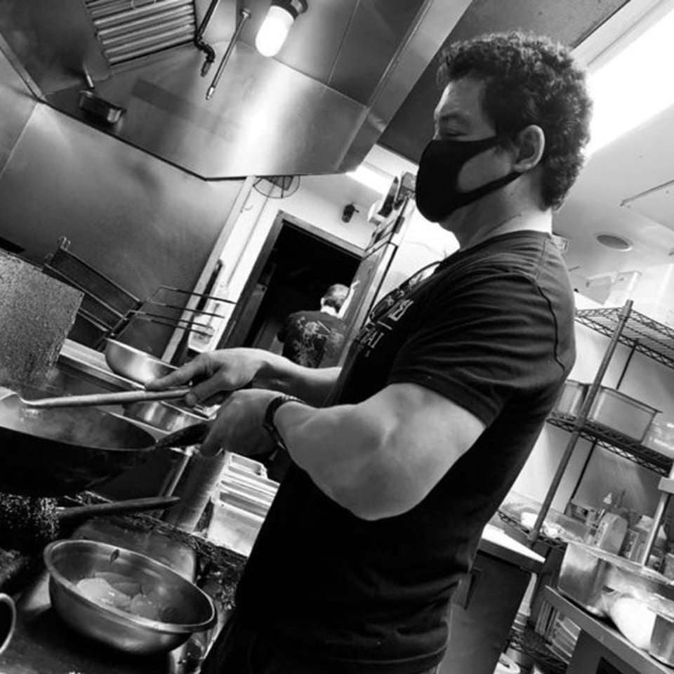A chef in a face mask cooks Thai food in a wok in a restaurant kitchen.