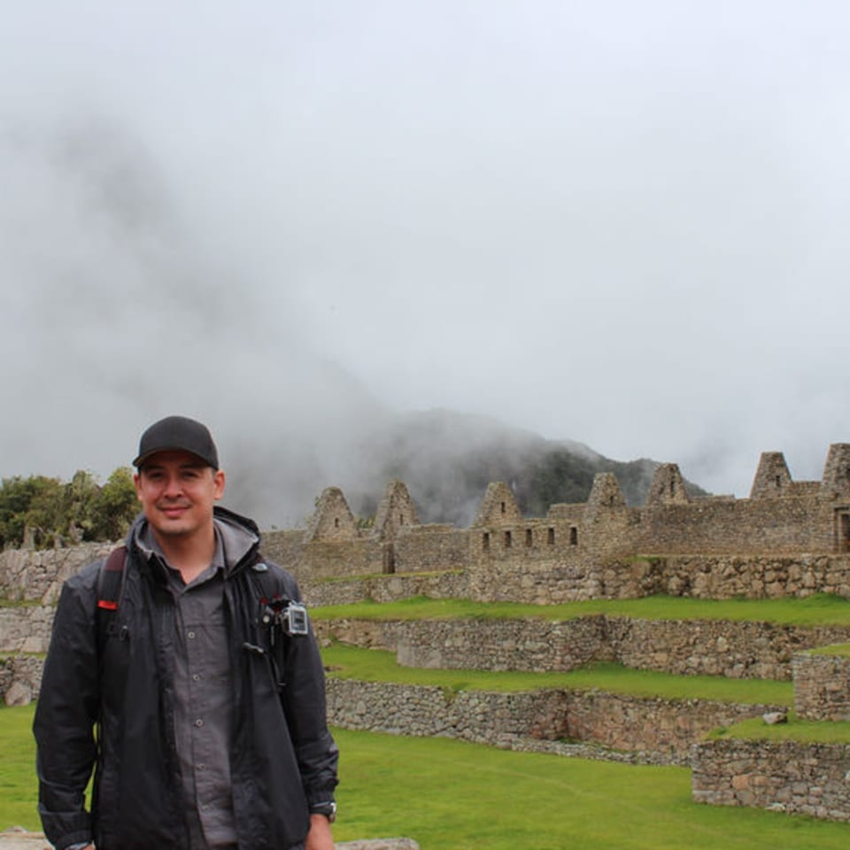 Man in a black jacket and cap smiling in front of ancient stone ruins and misty mountains.