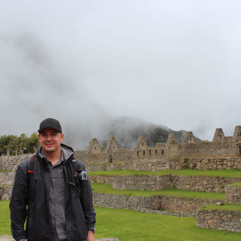 Man in a black jacket and cap smiling in front of ancient stone ruins and misty mountains.