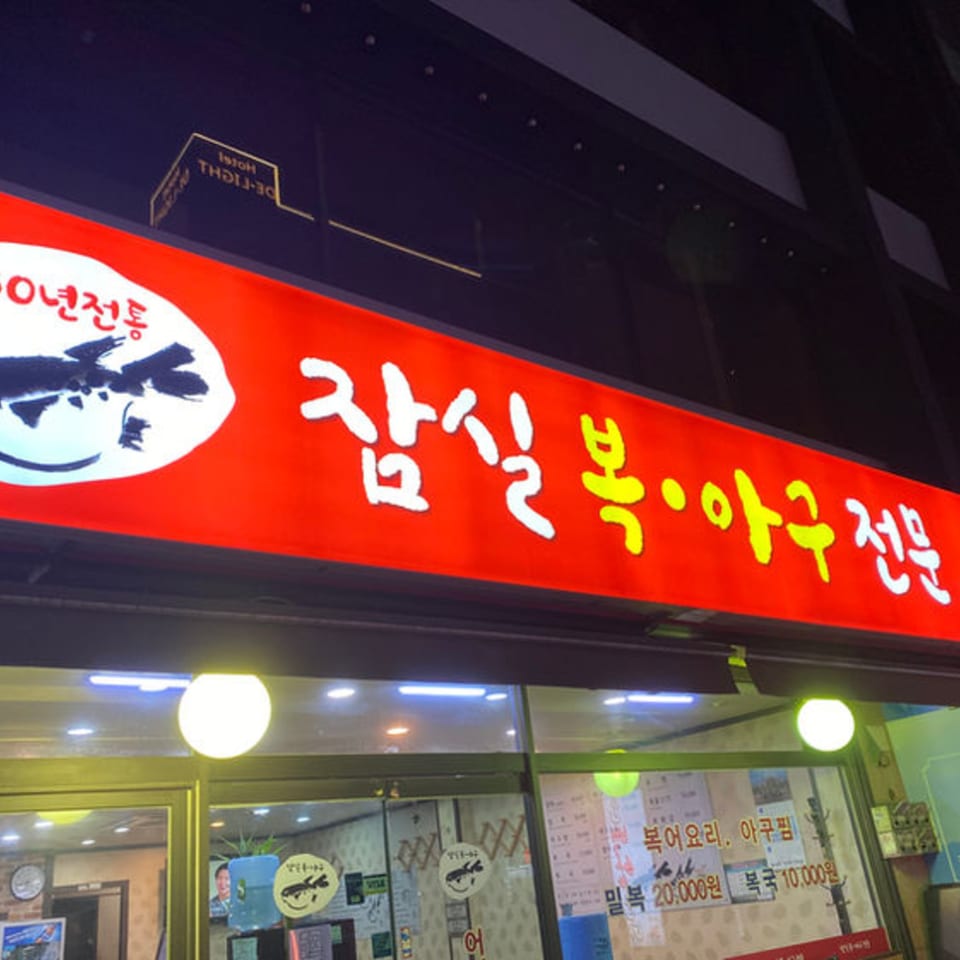 A bright red sign with Korean text in white and yellow, above a restaurant entrance at night.