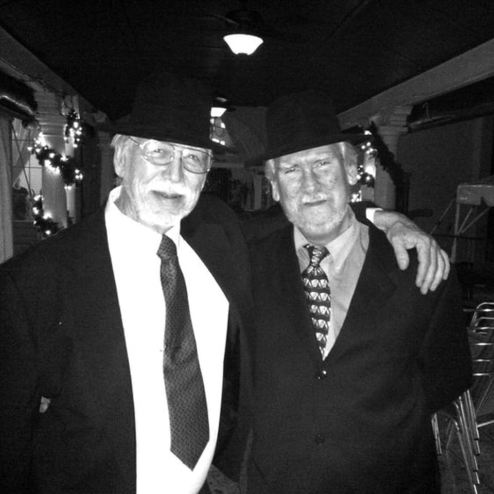Two older men in hats and suits, one with arm around the other, smiling in a dimly lit room.