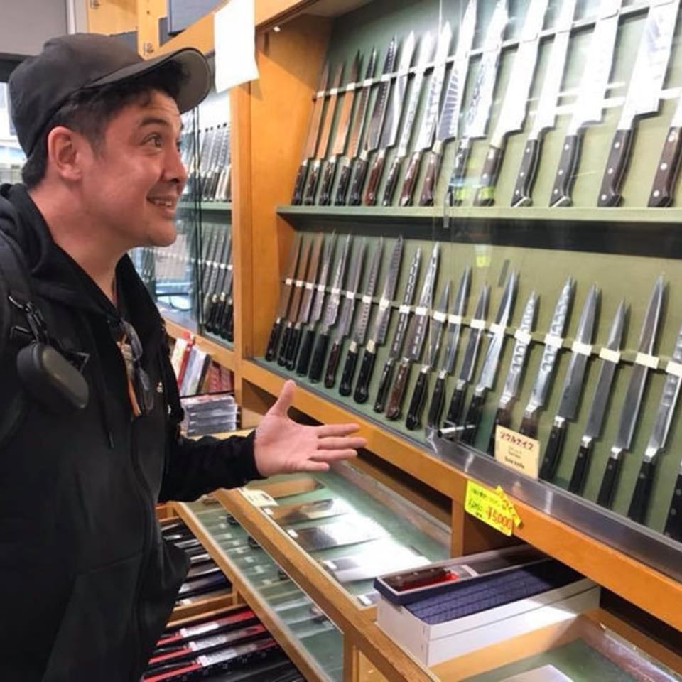 Man in a black cap and hoodie gesturing towards a display of knives in a store.