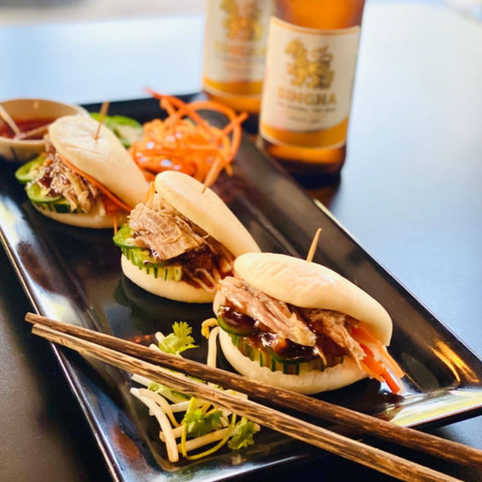 Three Thai bao buns with meat and vegetables, chopsticks, and two Singha beers on a black plate.
