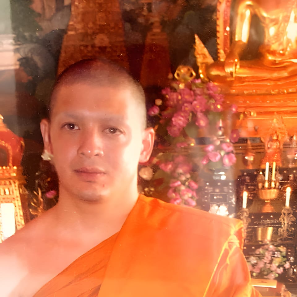 A man in an orange robe, possibly a monk, with a golden Buddha statue and pink flowers in the background.