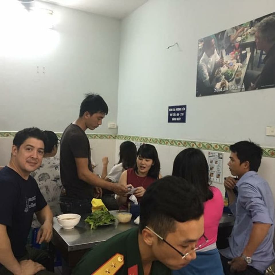 Diners seated at metal tables in a brightly lit, casual Thai restaurant with a poster on the wall.