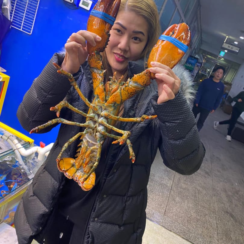 A woman in a black coat holds up a large, live orange lobster with blue bands on its claws.
