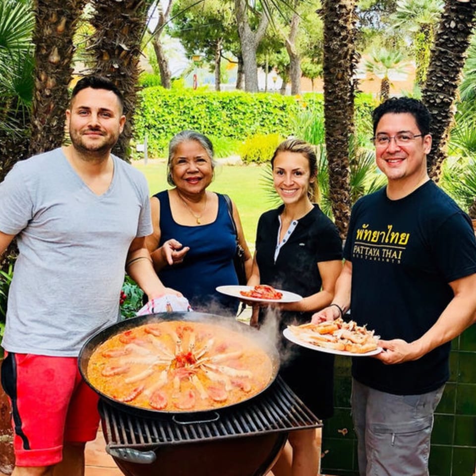 Four people smiling around a large paella pan cooking on a grill outdoors, with palm trees.