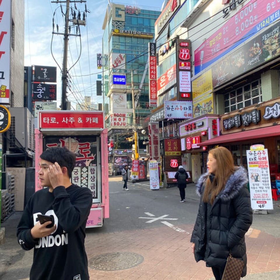 Two people walk on a busy street lined with shops and signs in an urban setting.