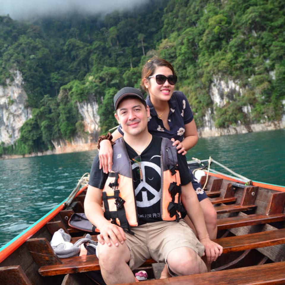 A couple in life vests smiles from a wooden boat on a lake surrounded by lush green mountains.