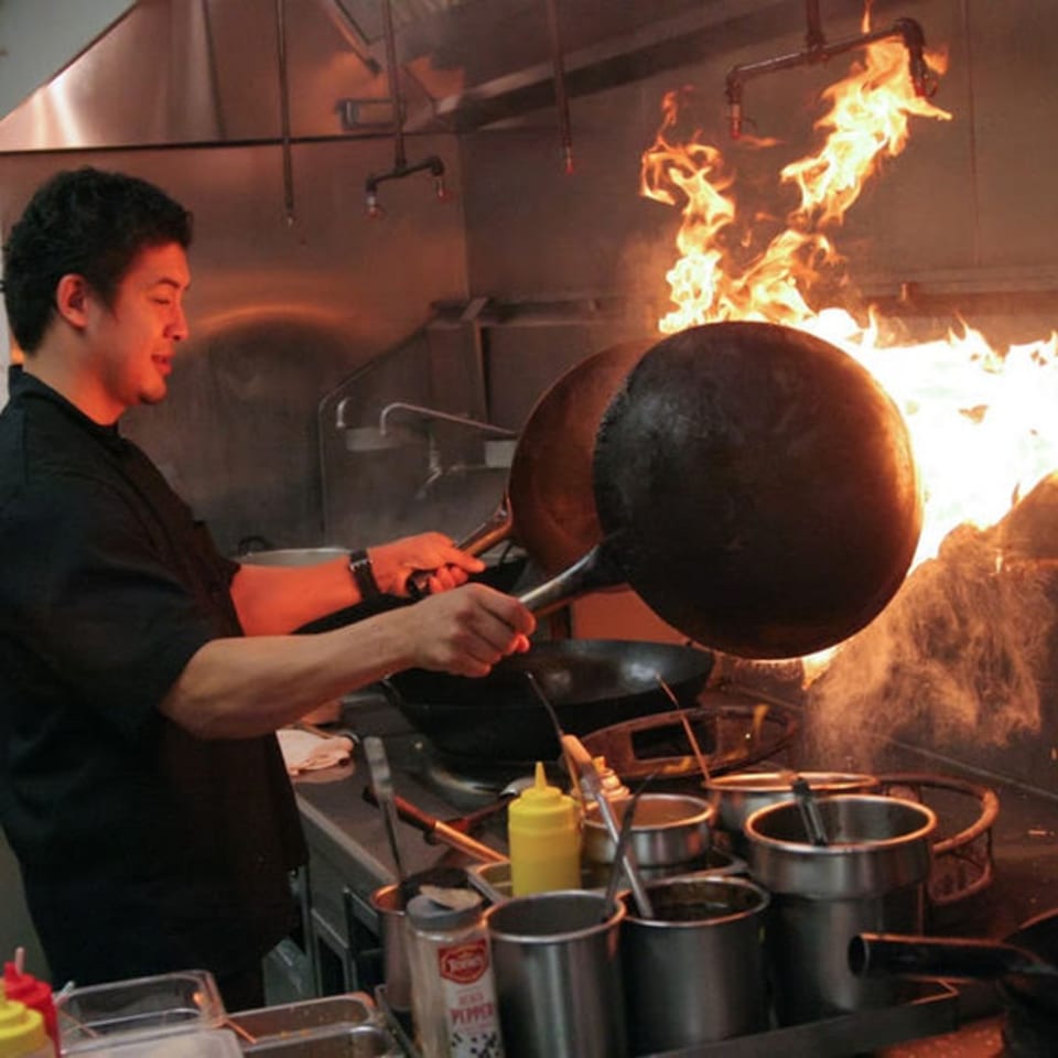A Pattaya Thai chef stir-frying with two woks, creating large flames in a busy kitchen.