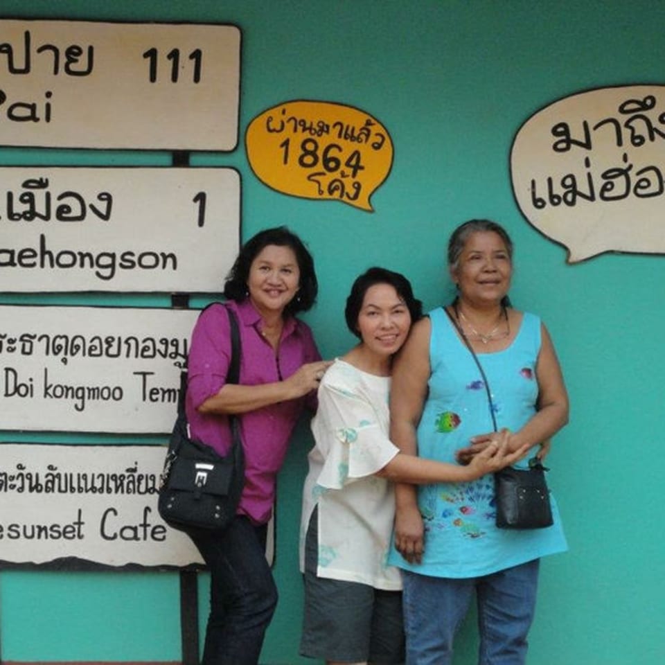 Three smiling Thai women pose in front of a teal wall with Thai signs.