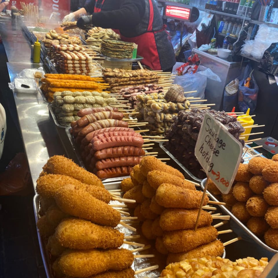 A vibrant street food stall displays various skewers of sausages, fried items, and other snacks.